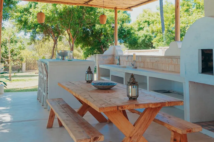 Built-in outdoor kitchen under a reed pergola: a rendered counter with a sink and tap, a wood-fired oven, a bar counter and a long wooden table with benches