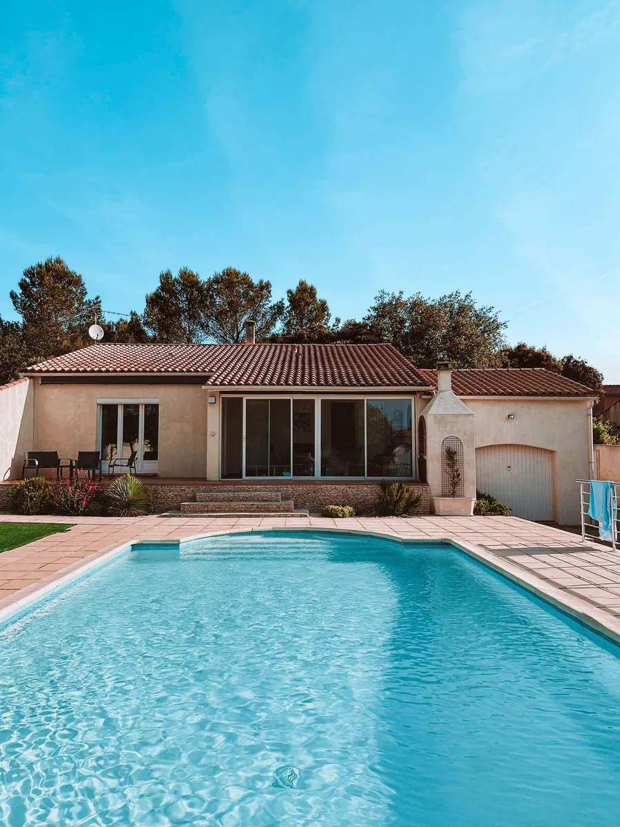 Rendered single-storey villa with a terracotta tile roof behind a kidney-shaped pool and a tiled deck