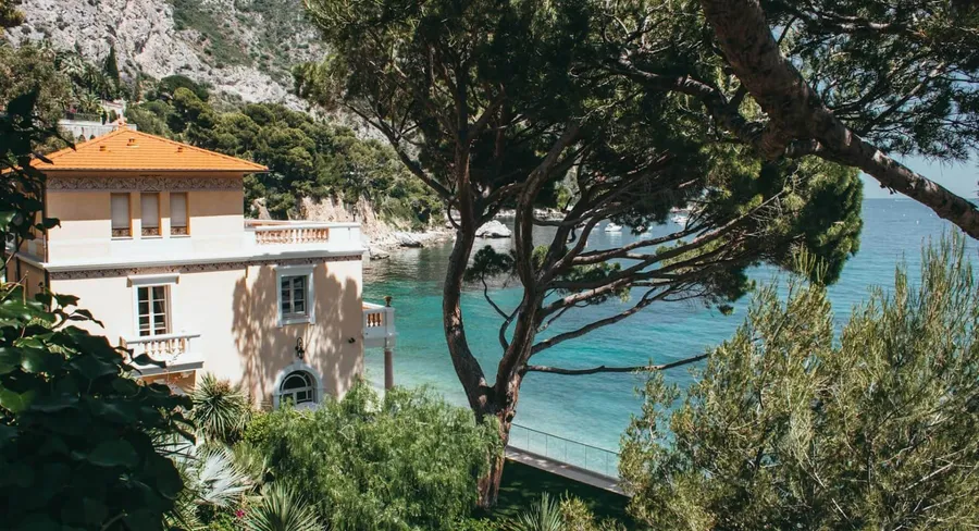 Rendered villa with a terracotta roof and balustraded terrace above a bay on the Riviera, framed by umbrella pines