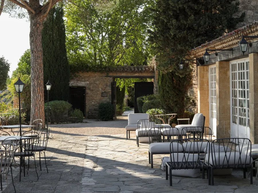 Stone-paved terrace with cushioned loungers beside an old stone farmhouse and umbrella pines in Provence