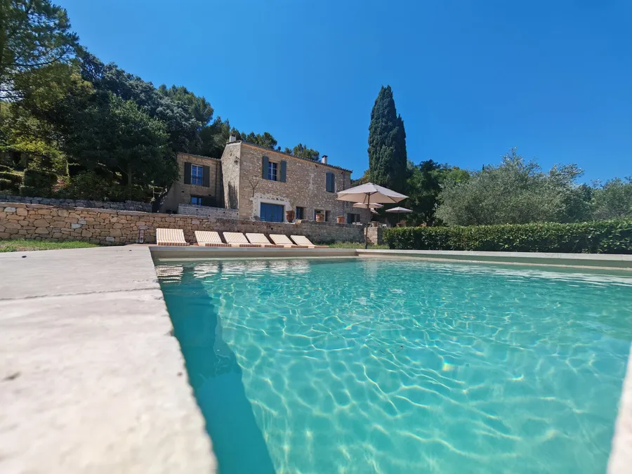 Restored stone house with grey shutters above a swimming pool, cypress and olive trees behind