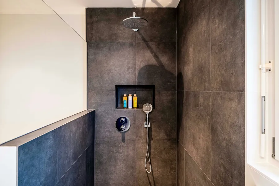 Walk-in shower in dark stone-look tiles with a low tiled side wall, a recessed niche holding bottles, a ceiling rain head and a hand shower on a hose